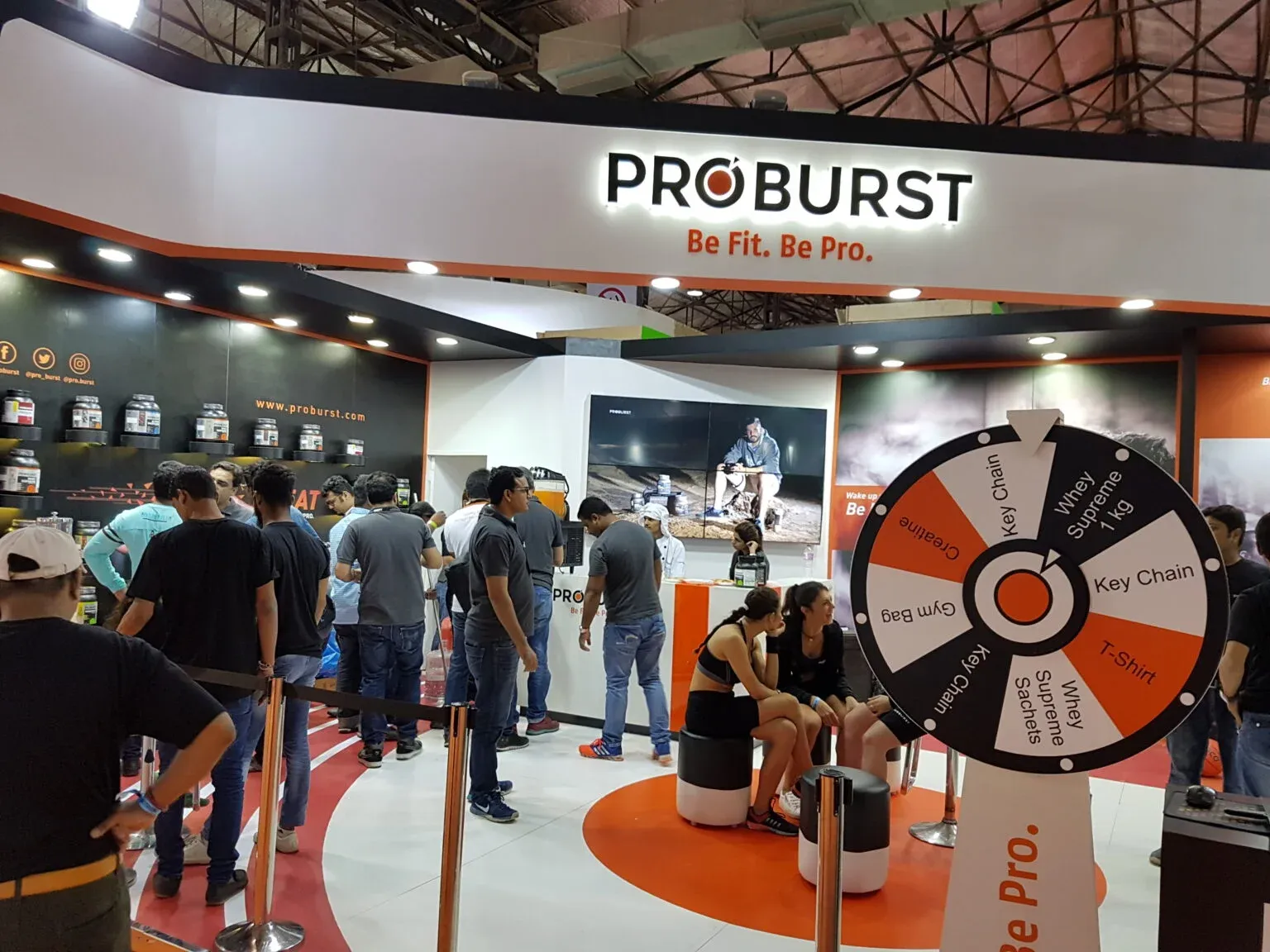 Crowded exhibition hall with visitors around a branded display and a prize wheel.
