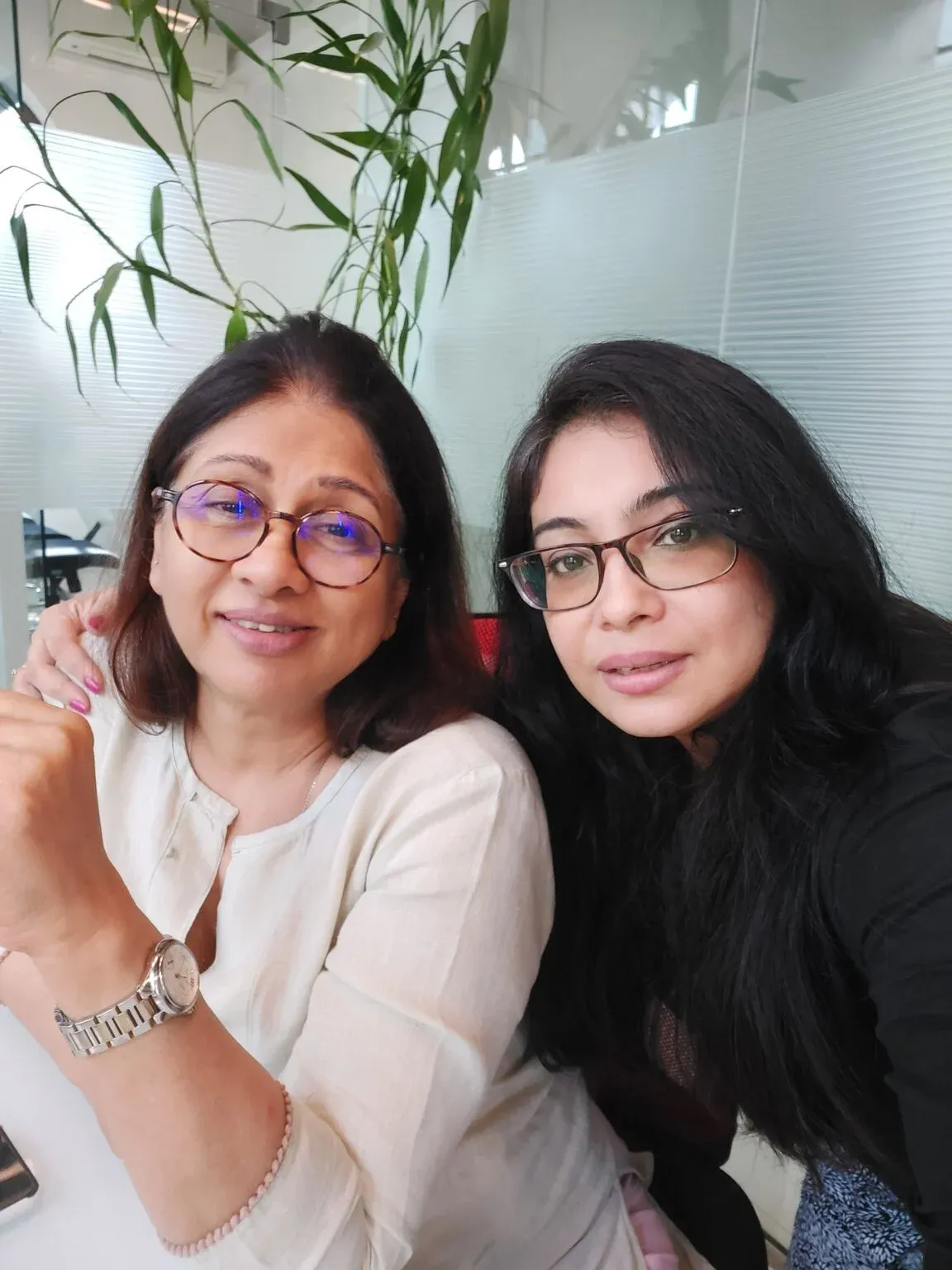 Two women smiling together in a bright office with frosted glass and plants behind them.