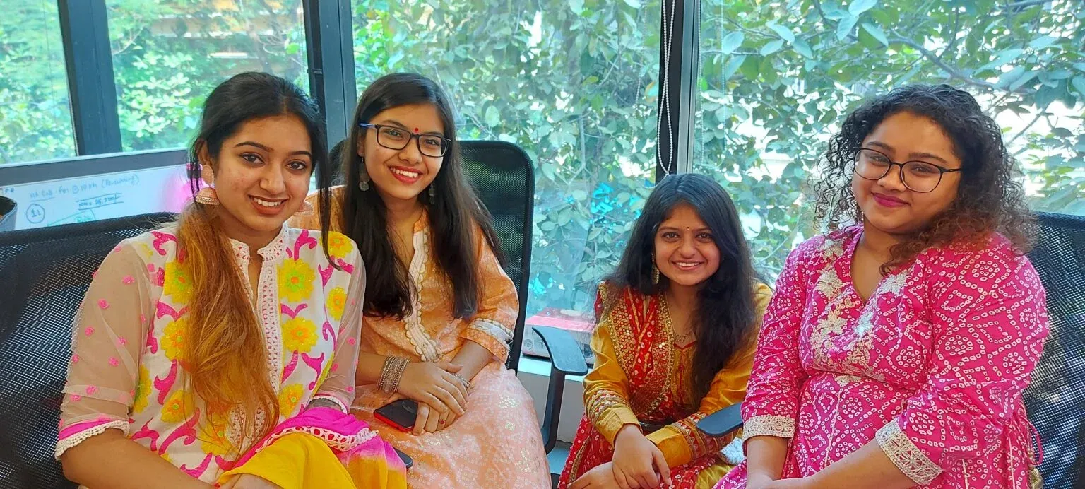 Four women in colourful festive attire smiling together in office chairs by a window.