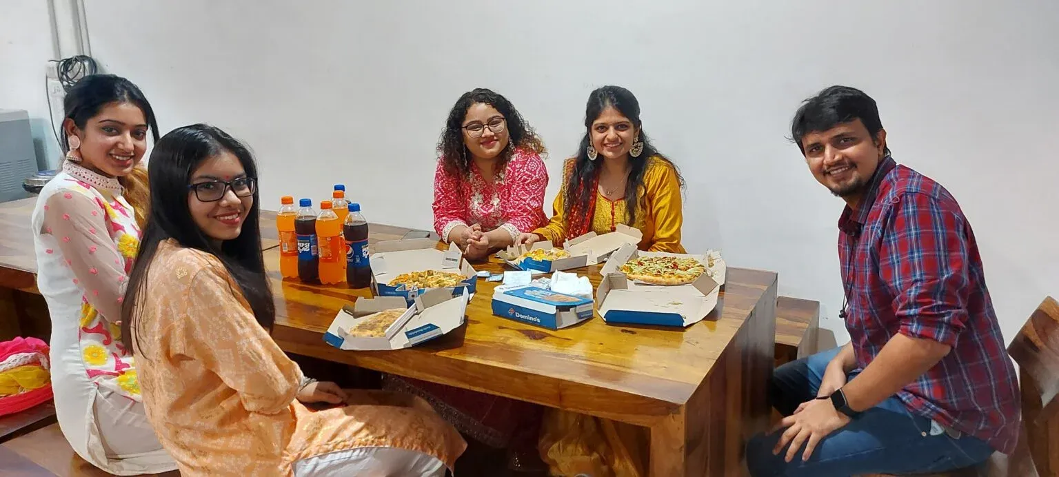 Five colleagues sharing pizza and soft drinks around a wooden table indoors.
