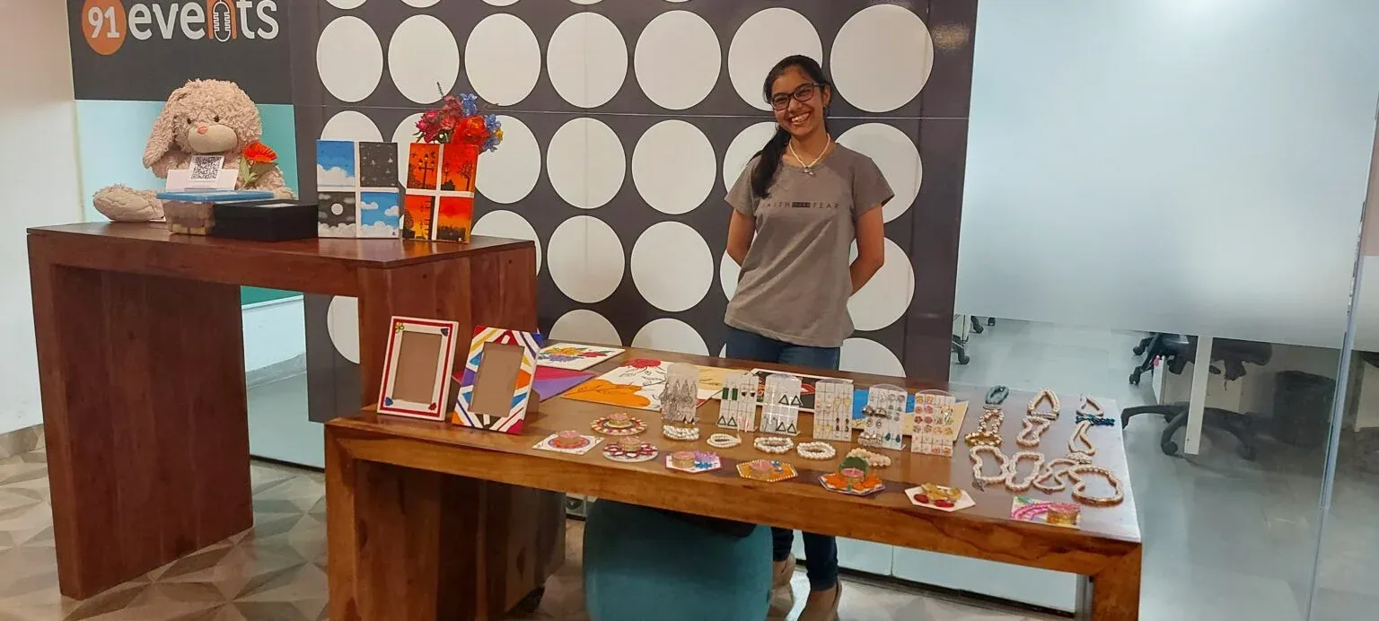 Smiling woman standing behind a wooden table of handmade jewellery and small paintings.