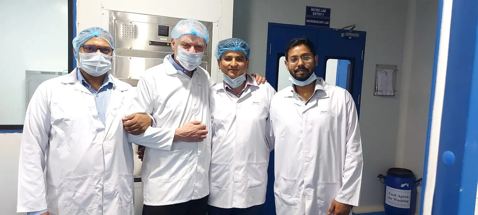 Four people in lab coats and hairnets standing together inside a laboratory facility.