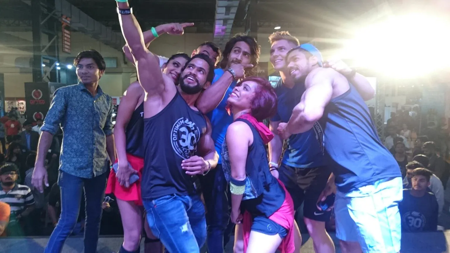 Group of people posing for a selfie on a raised stage above a crowded hall.