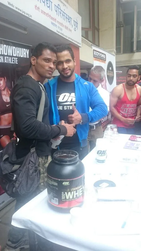 Two men posing behind a table of sports nutrition products at a public fitness event.