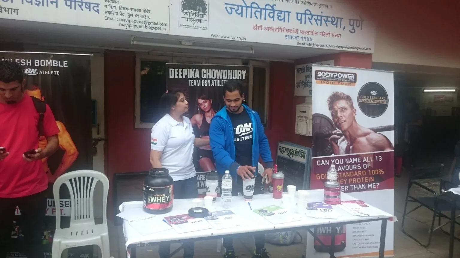 Two people behind a product display table with tall banners at an outdoor public event.