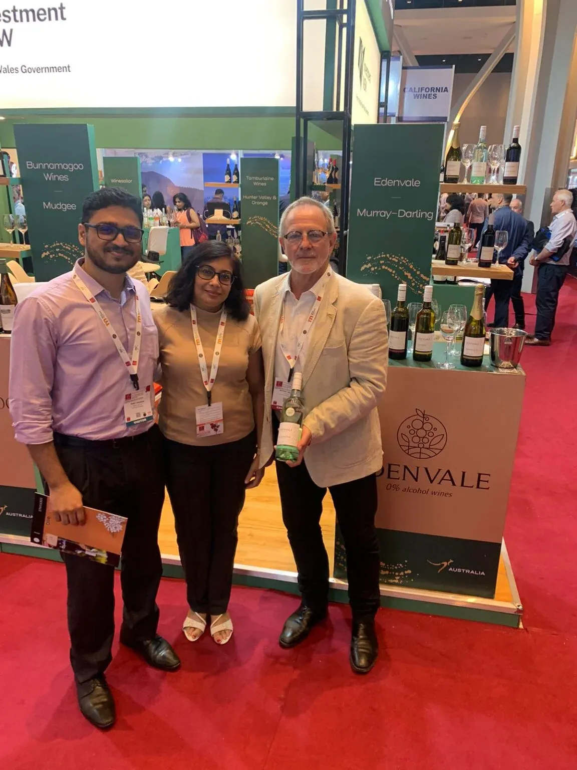Three people with event lanyards posing in front of a wine display at a trade exhibition.