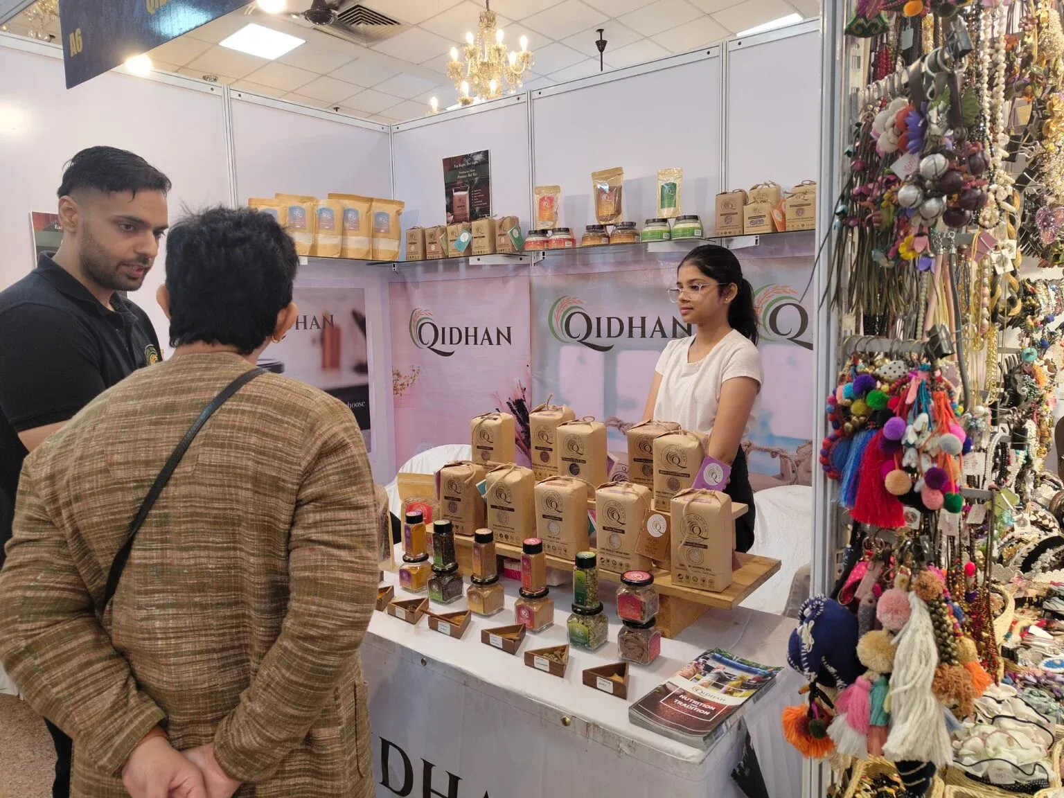 Woman behind a counter of packaged goods and sample jars speaking with visitors at an exhibition.