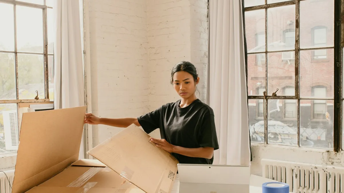 A shop owner opening a cardboard shipping box at a table beside a laptop