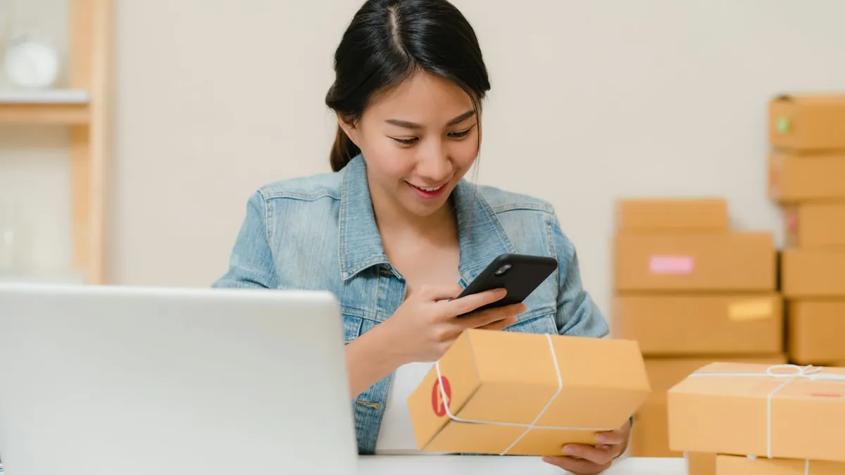 A small-business owner scanning a parcel with a phone at a desk surrounded by boxes